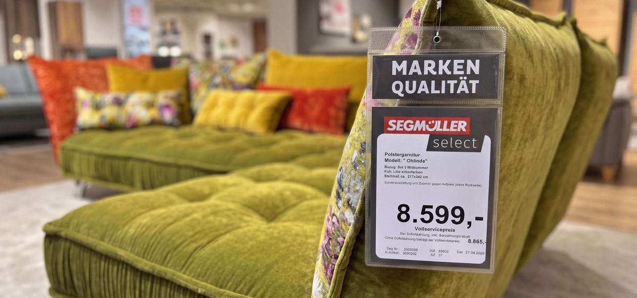 Ein grünes Samtsofa mit einem Preisschild von 8.599 Euro in einem Ausstellungsraum, im Hintergrund bunte Kissen und ein Holzboden.