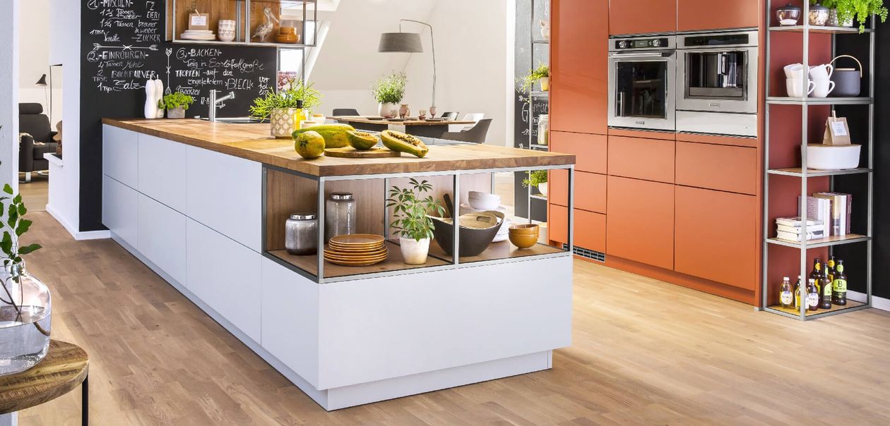 Moderne Küche mit weißer Kochinsel, Holzarbeitsplatte und Pflanzen. Im Hintergrund stehen orangefarbene Schränke mit eingebauten Backöfen.
