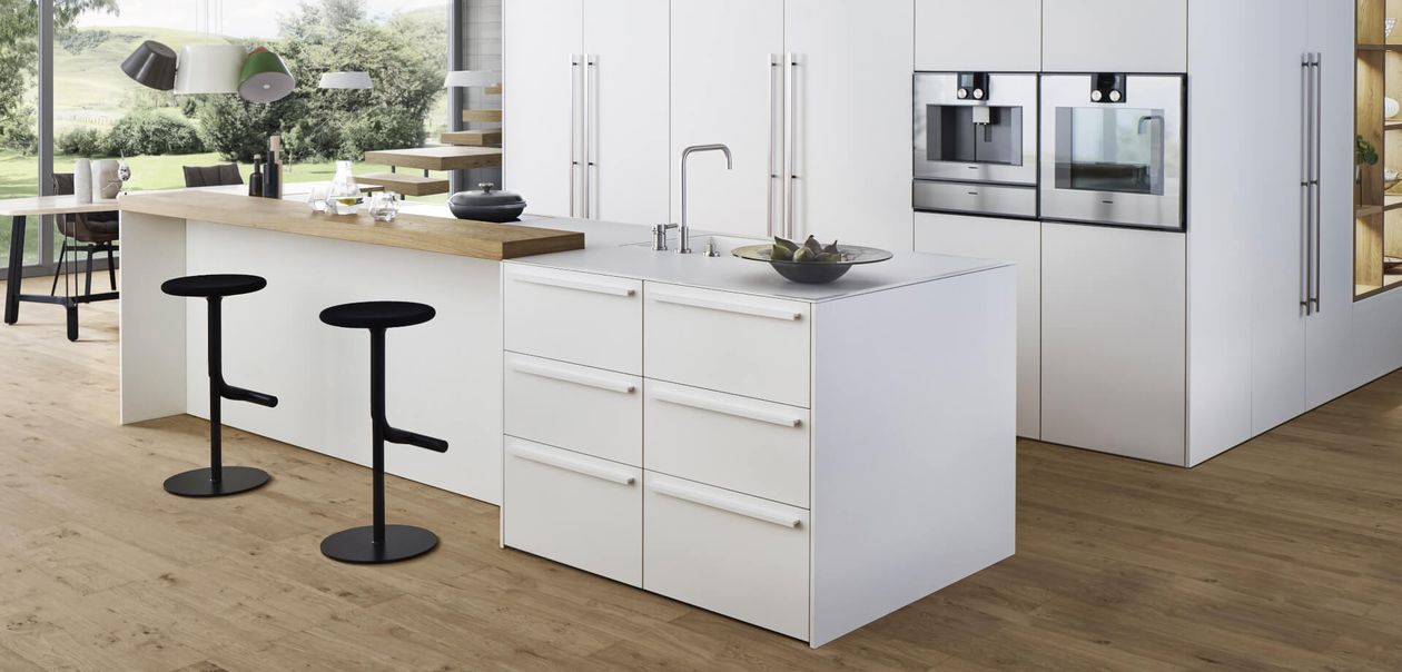 Moderne Küche mit weißen Schränken, einer Holzinsel mit zwei schwarzen Hockern, Einbauöfen und großen Fenstern mit Blick ins Grüne.