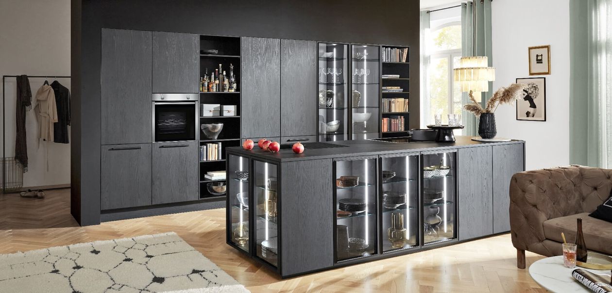 Moderne Küche mit dunklen Schränken, einer Insel mit Glasfront, auf der Geschirr ausgestellt ist, und Äpfeln auf der Arbeitsplatte. Durch ein großes Fenster fällt helles Licht herein.