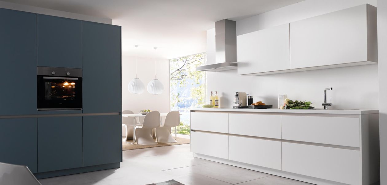 Moderne Küche mit eleganten weißen Schränken, Einbauherd und einem Essbereich mit großen Fenstern und Pendelleuchten im Hintergrund.