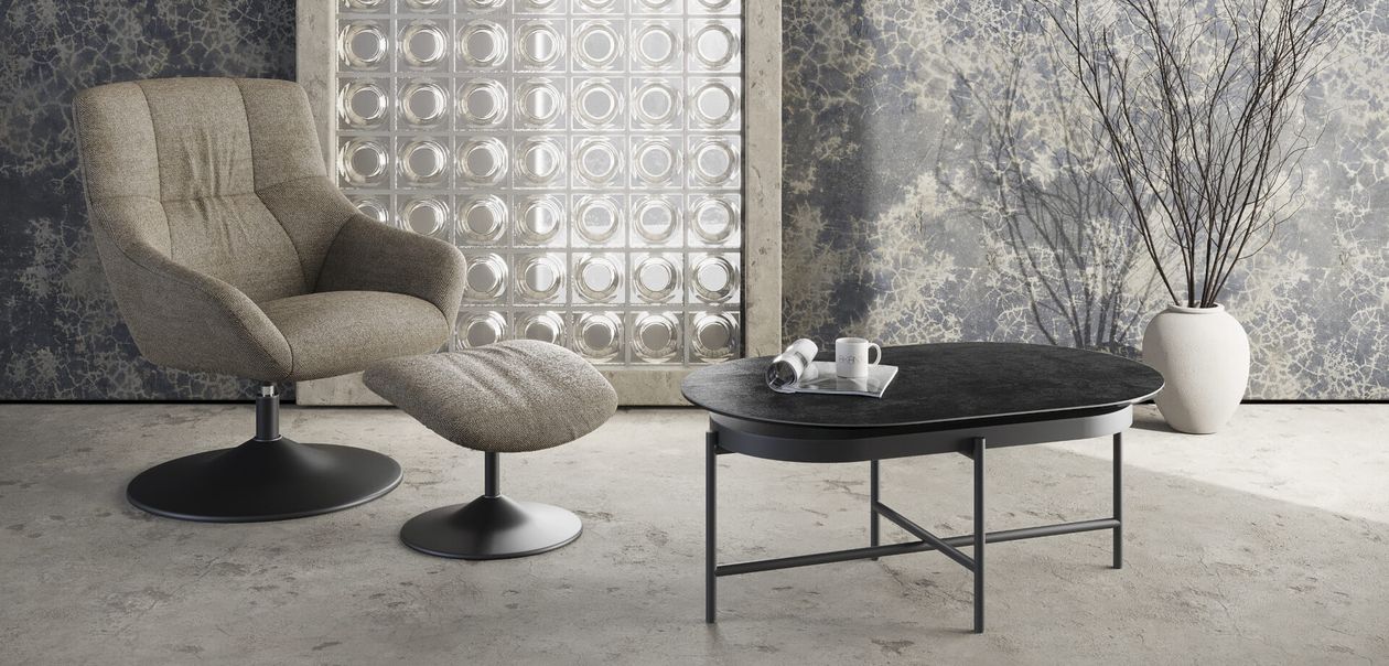 Modernes Wohnzimmer mit grauem Sessel, passendem Ottoman, ovalem Couchtisch, strukturierter Tapete, Glasbausteinfenster und Vase mit Zweigen.