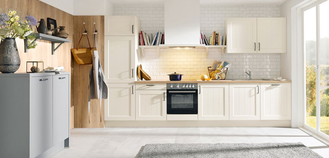 Moderne Küche mit weißen Schränken, einem Einbauofen und einer Arbeitsplatte aus Holz. Auf Regalen stehen Bücher und Dekoartikel. Durch ein großes Fenster fällt natürliches Licht herein.
