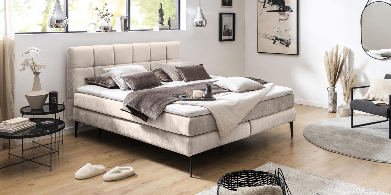 Modernes Schlafzimmer mit einem beigen Polsterbett, gemütlichen Kissen und Überwürfen, ergänzt durch stilvolle Dekoration und ein großes Fenster. Neutrale Farbpalette.