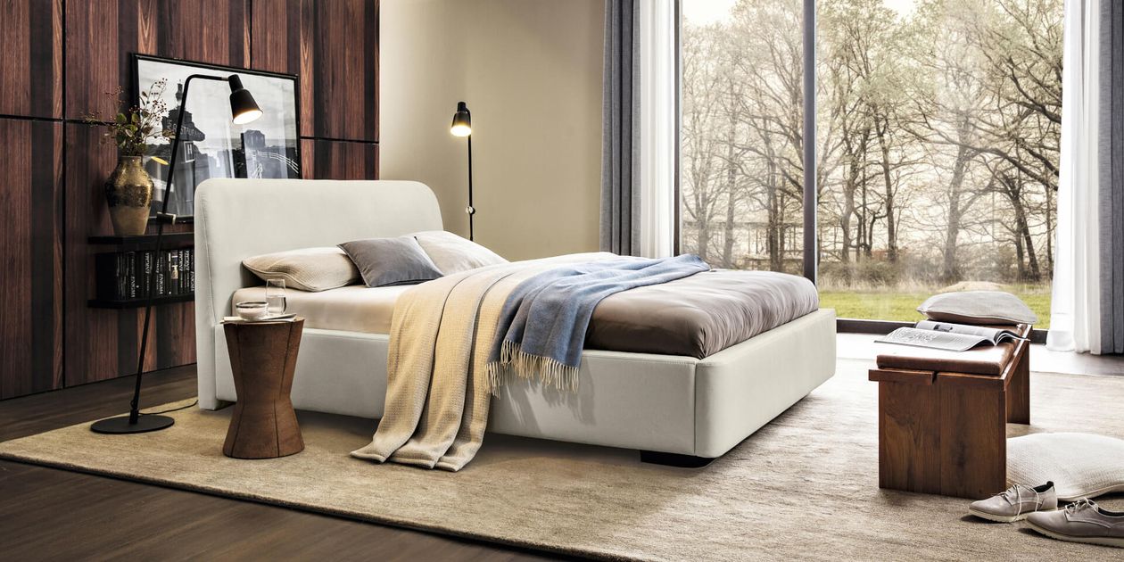 Modernes Schlafzimmer mit einem weißen Bett, braunen und blauen Decken, Holzakzenten und großen Fenstern mit Blick auf Bäume. Sanfte Beleuchtung schafft eine gemütliche Atmosphäre.