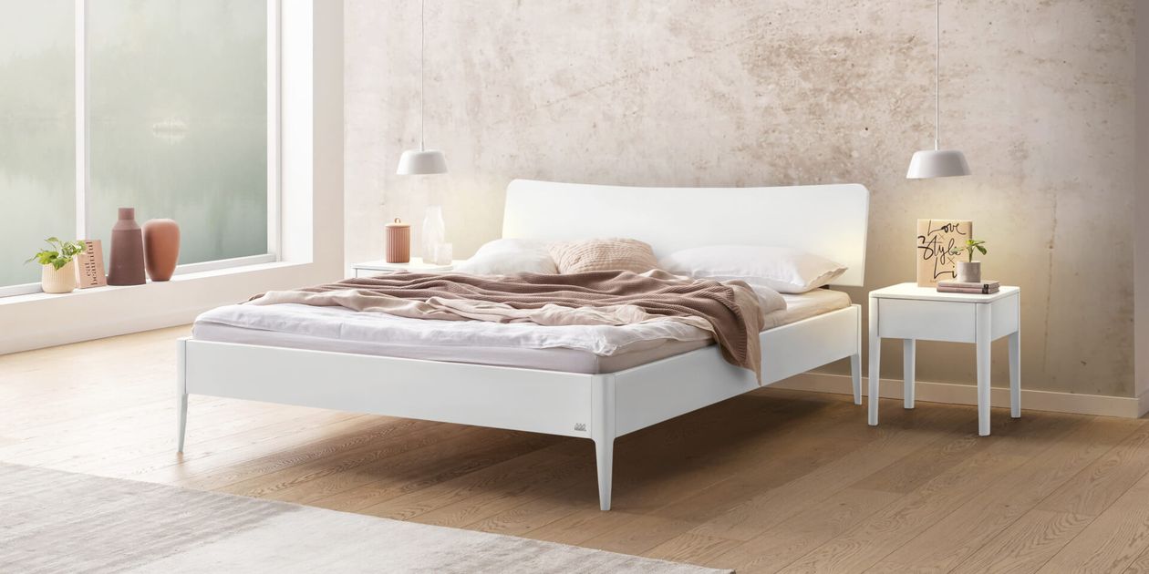 Ein minimalistisches Schlafzimmer mit einem weißen Bett und unordentlicher Bettwäsche, flankiert von zwei Nachttischen mit Lampen, vor einer strukturierten Wand und einem Hartholzboden.