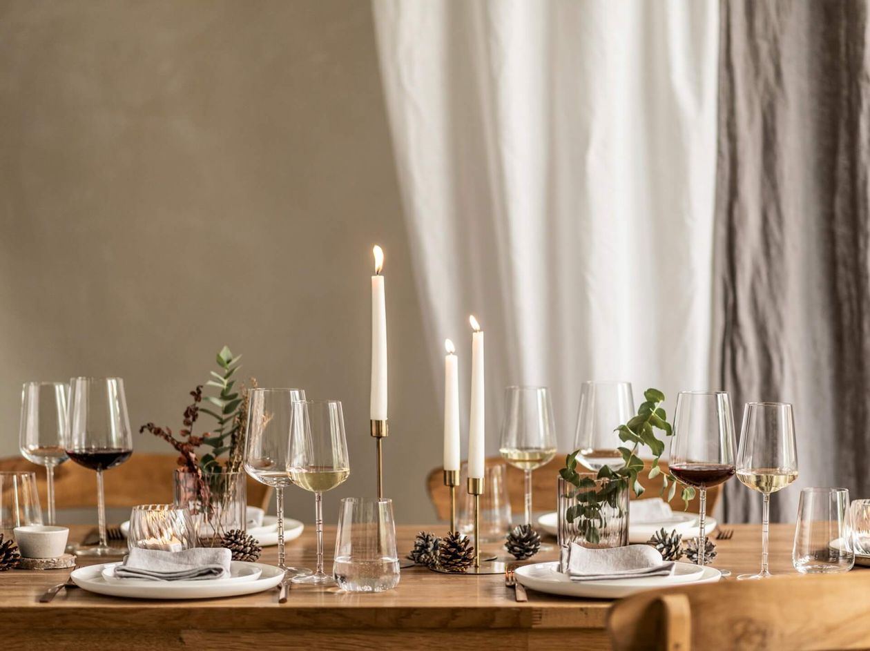 Elegante Tischdekoration mit Weingläsern, brennenden Kerzen und Tannenzapfendekorationen auf einem Holztisch vor neutralem Hintergrund.