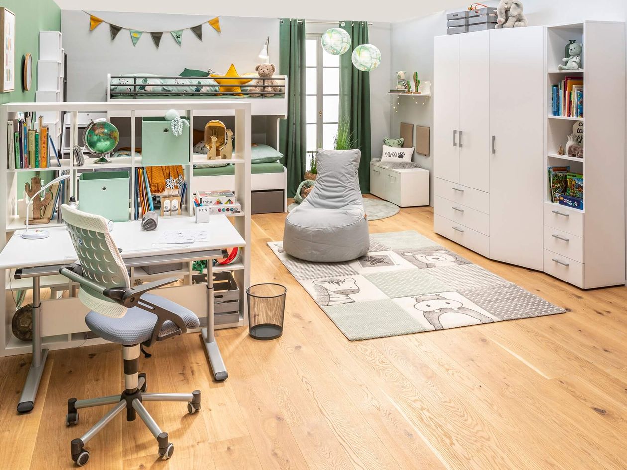 Helles Kinderzimmer mit weißem Hochbett, darunter integriertem Schreibtisch mit ergonomischem Drehstuhl, weißem Kleiderschrank und Bücherregal. Der Raum ist in Grün- und Grautönen gestaltet, mit bunter Wimpelkette, grünen Vorhängen, kugelförmigen Hängeleuchten in Weltkarten-Optik und einem Teppich mit Tiermotiven. Ein grauer Sitzsack steht in der Raummitte auf dem Eichenparkett.