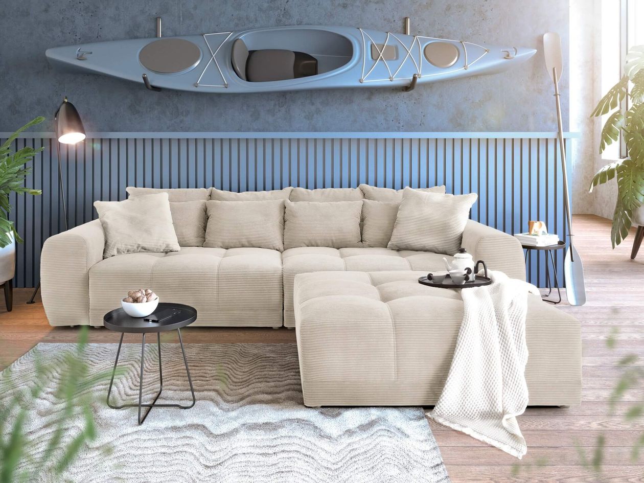 Großes cremefarbenes Cord-Sofa mit Hocker in einem maritim eingerichteten Wohnzimmer. An der Wand hängt ein graues Kajak, darunter eine blaue Holzlamellen-Verkleidung. Ein grau-weißer Wellenteppich und Beistelltische ergänzen das Ambiente.