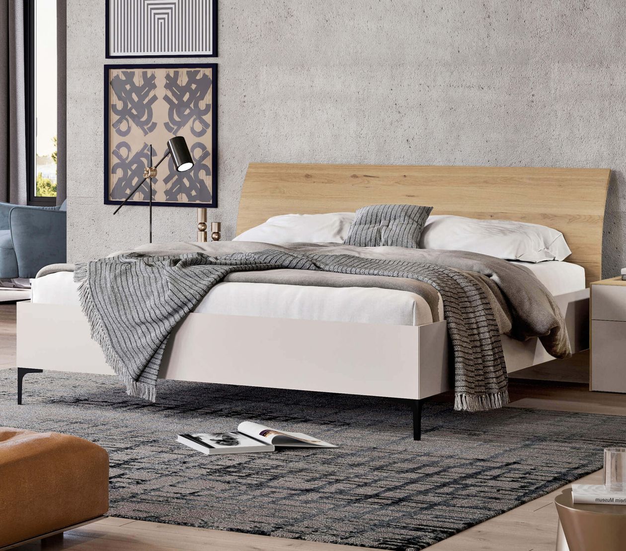 Modernes Schlafzimmer mit Doppelbett, grauen Decken und Holz-Kopfteil. Kunstwerke an der Betonwand, schwarze Lampe, offenes Buch auf Teppich.