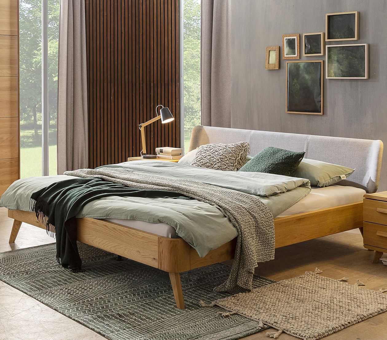 Modernes Schlafzimmer mit Holzbett, grüner Bettwäsche und Kissen. Neben dem Bett steht eine Lampe auf einem Nachttisch. Kunst an der Wand.