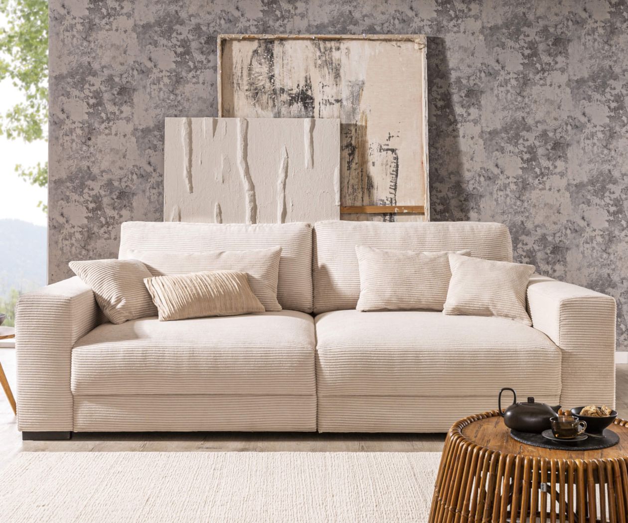 Beige Sofa mit Kissen vor bemalter Wand. Zwei abstrakte Bilder lehnen an der Wand. Rattantisch mit Teekanne und Schale im Vordergrund.