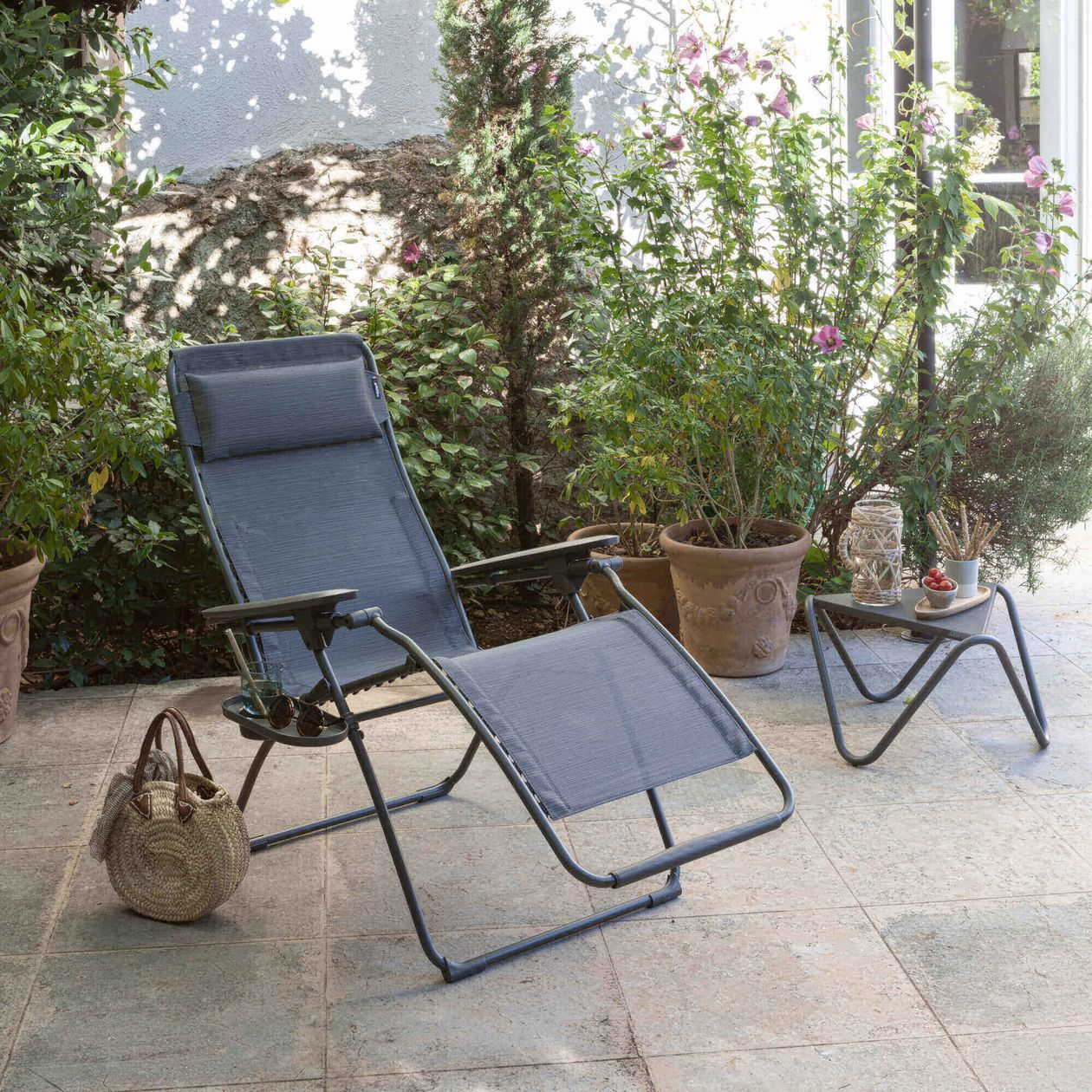 Liegestuhl und kleiner Tisch mit Getränken auf einer Terrasse, umgeben von blühenden Pflanzen in Töpfen, bei sonnigem Wetter.