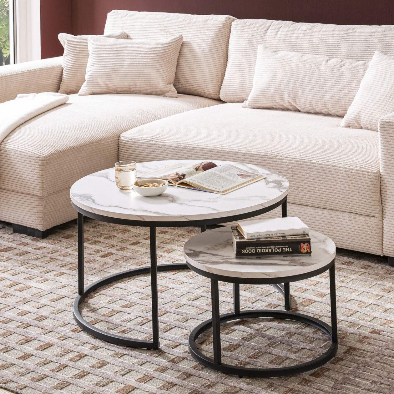 Ein modernes Wohnzimmer mit einem beigen Ecksofa, Marmor-Stapeltischen mit Büchern, Getränken und Snacks auf einem karierten Teppich.