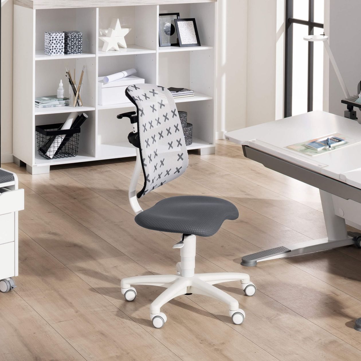 Moderner Bürostuhl mit weißem Gestell und grauem Sitz daneben ein weißer Schreibtisch und ein Regal mit Dekoartikeln auf Holzfußboden.
