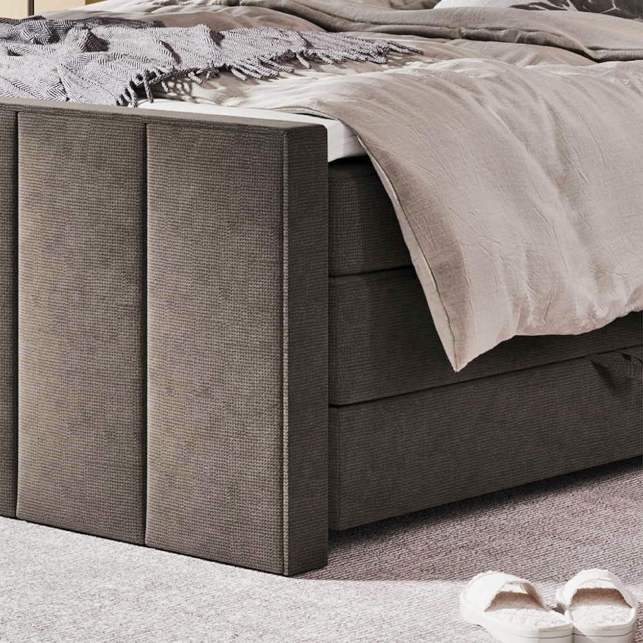 Braunes Boxspringbett mit beigefarbener Bettdecke und grauer Decke, auf grauem Teppich. Weiße Hausschuhe am Boden.
