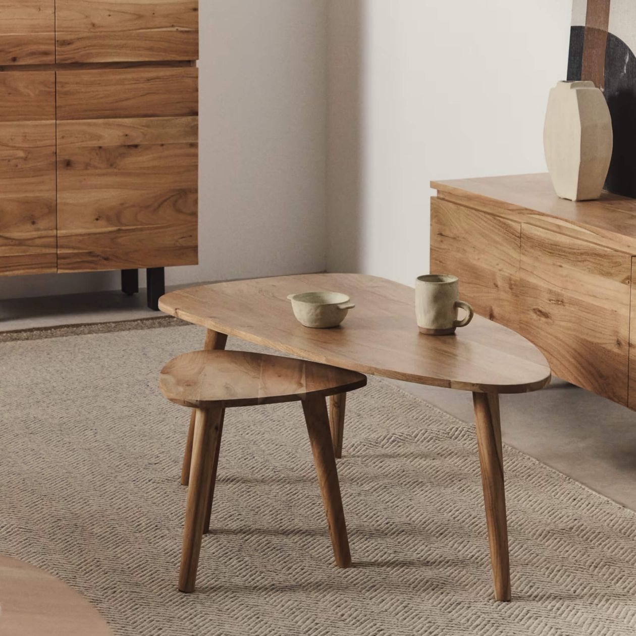 Ein Set aus ineinander stapelbaren Holztischen mit Tassen darauf, platziert in einem gemütlichen Raum mit passenden Holzmöbeln und einem strukturierten beigen Teppich.