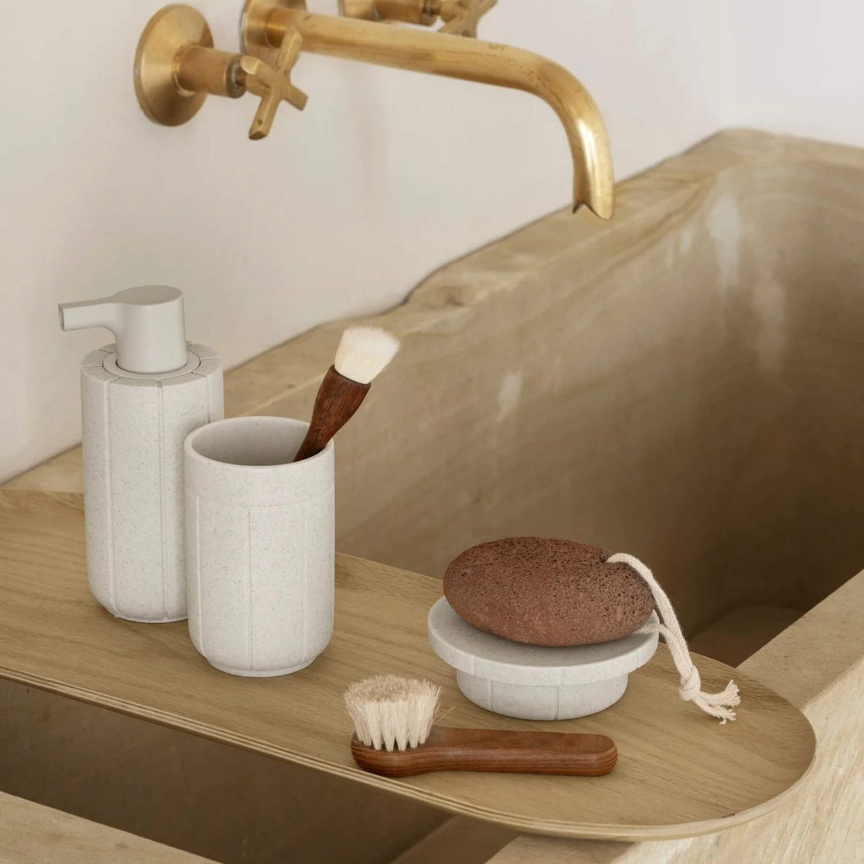 Natürliche Badezimmerdekoration: Seifenspender, Becher mit Pinsel, Bimsstein und Bürste auf Holzablage, vor Bronze-Wasserhahn.