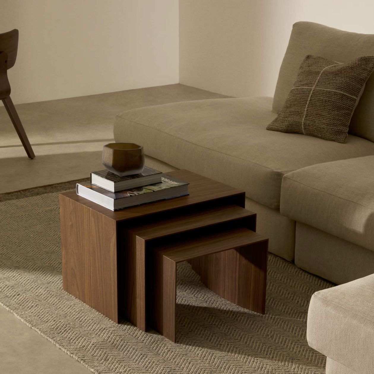 Ein gemütliches Wohnzimmer mit einem beigen Sofa, gemusterten Kissen, ineinander gestellten Beistelltischen aus Holz, Büchern und einer Schale darauf, alles auf einem strukturierten Teppich.