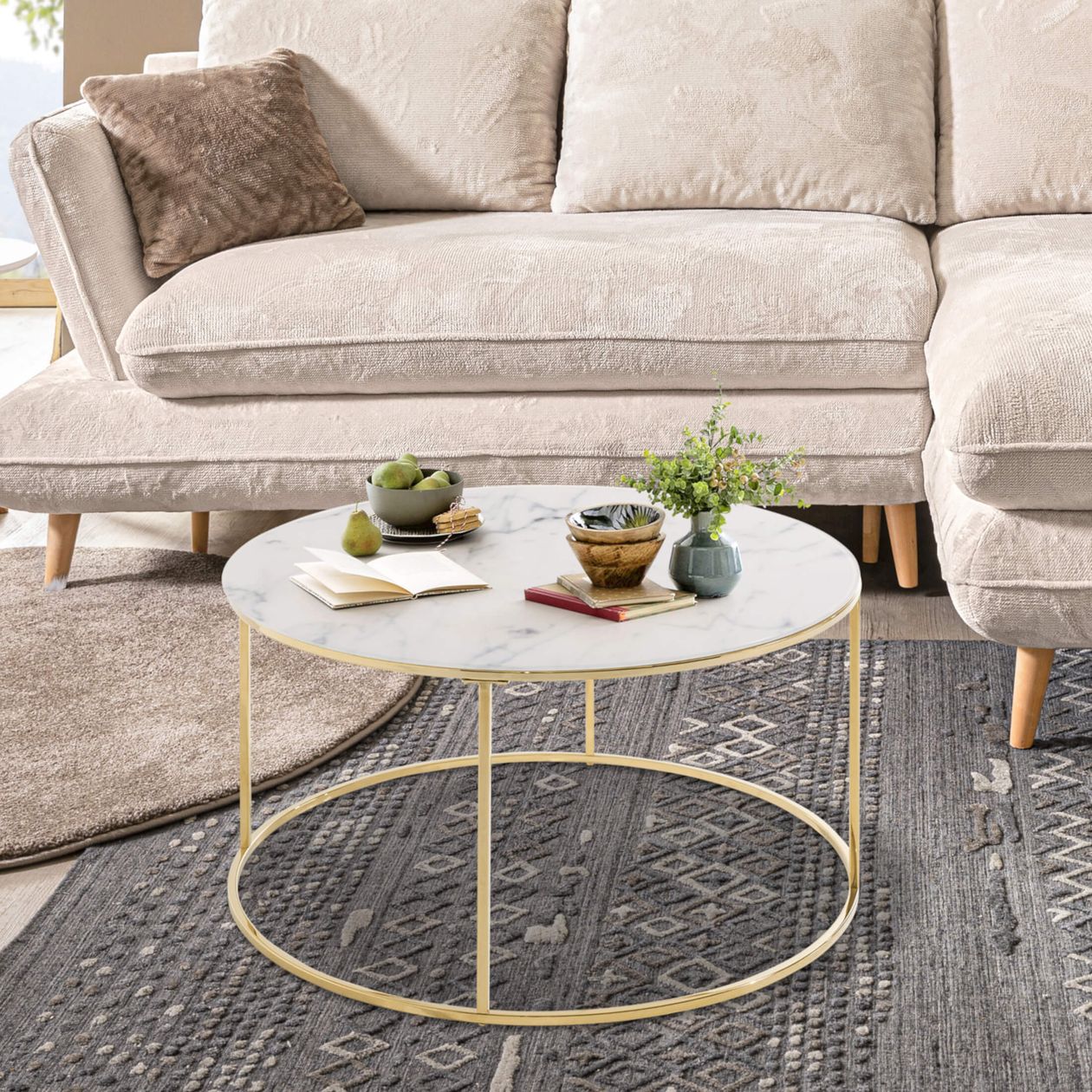 Ein elegantes Wohnzimmer mit einem beigen Sofa, gemusterten Kissen und einem runden Marmor-Couchtisch, auf dem Bücher, Geschirr und eine kleine Pflanze stehen.