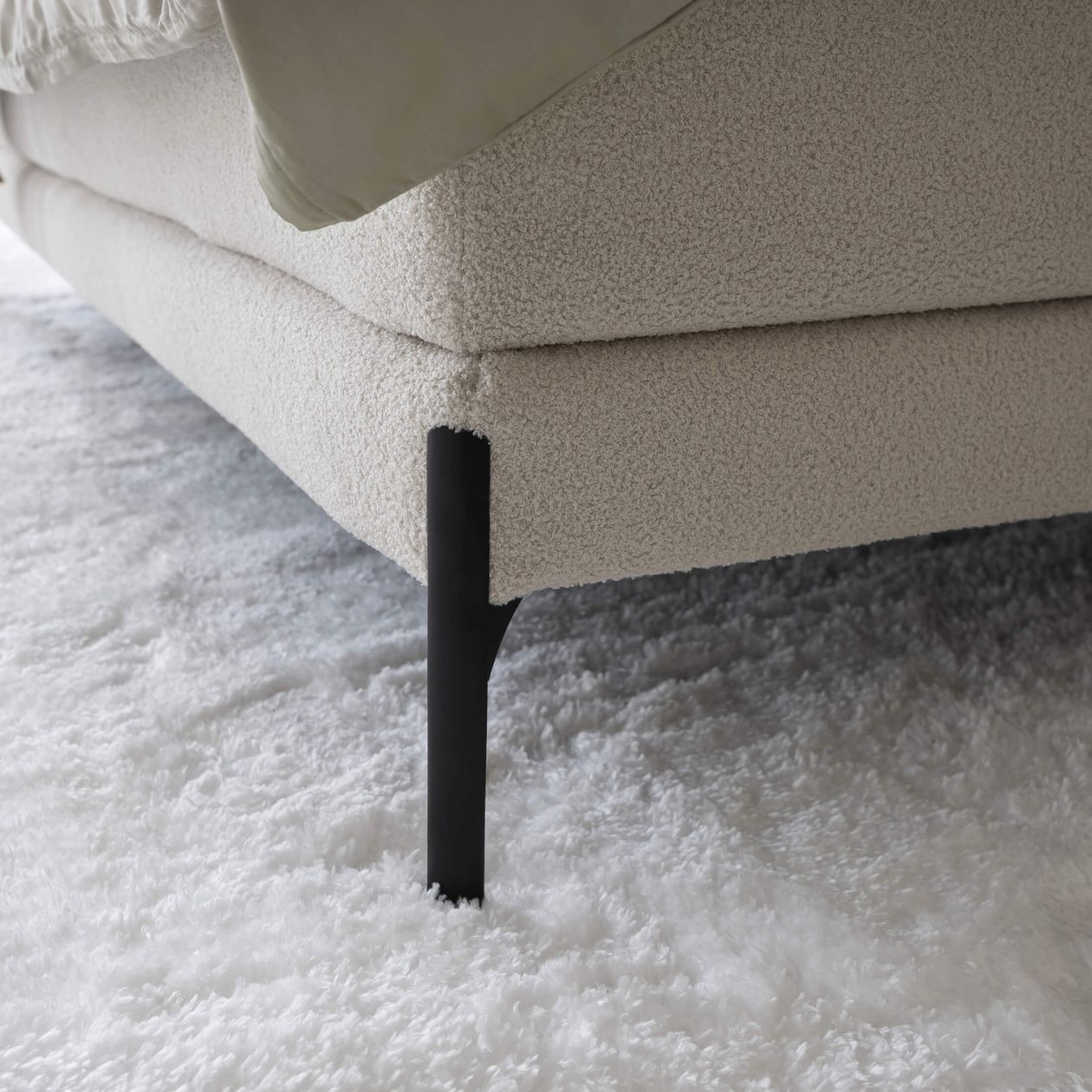 Nahaufnahme eines beigefarbenen Sofas mit schwarzem Metallbein auf einem flauschigen, weißen Teppichboden.