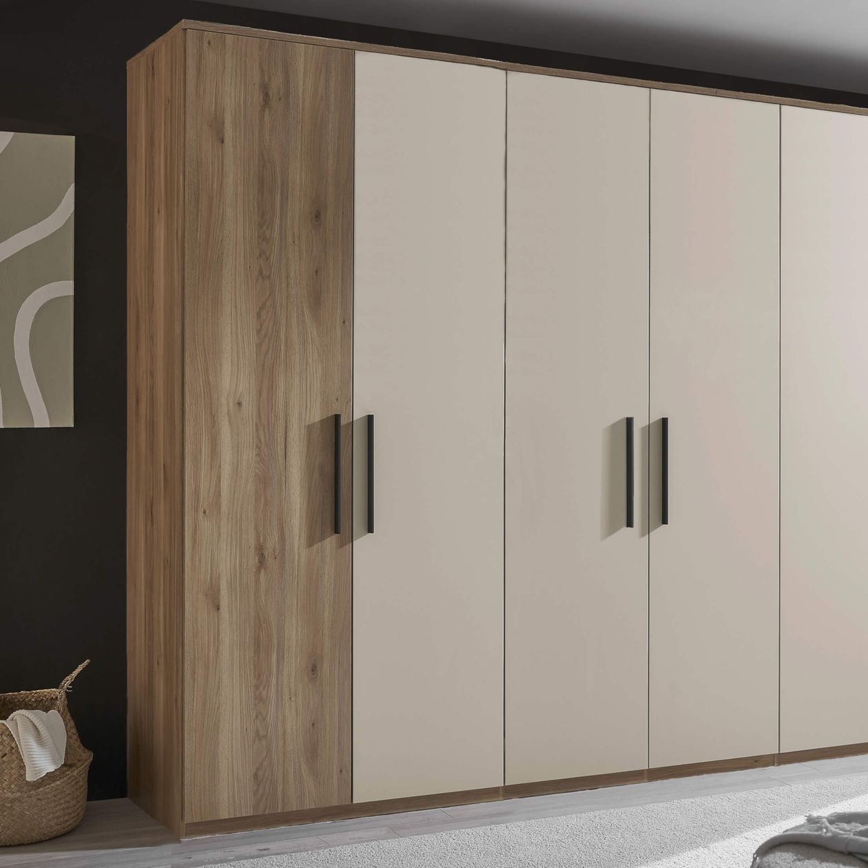 Großer Schrank mit Holzakzent und cremefarbenen Türen, schwarzen Griffen, steht auf Teppich neben einem Korb, weiße Wand mit Kunstwerk.