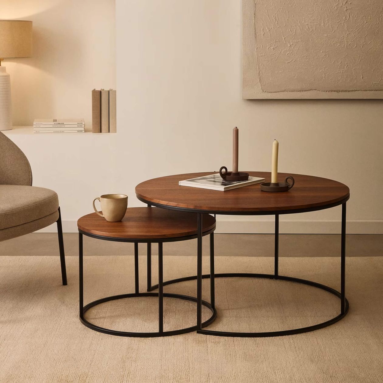 Zwei runde Couchtische aus Holz mit schwarzen Metallbeinen, die übereinander gestapelt sind, mit Kerzen, einem Buch und einer Tasse in einem minimalistisch eingerichteten Raum.
