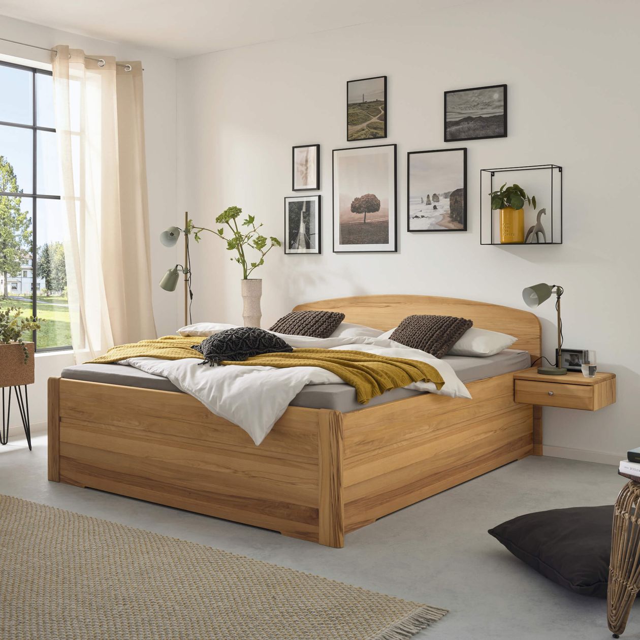 emütliches Schlafzimmer mit einem Holzbett, einer gelben Decke, Wandbildern, Topfpflanzen und einem großen Fenster mit transparenten Vorhängen, das den Blick ins Grüne freigibt.