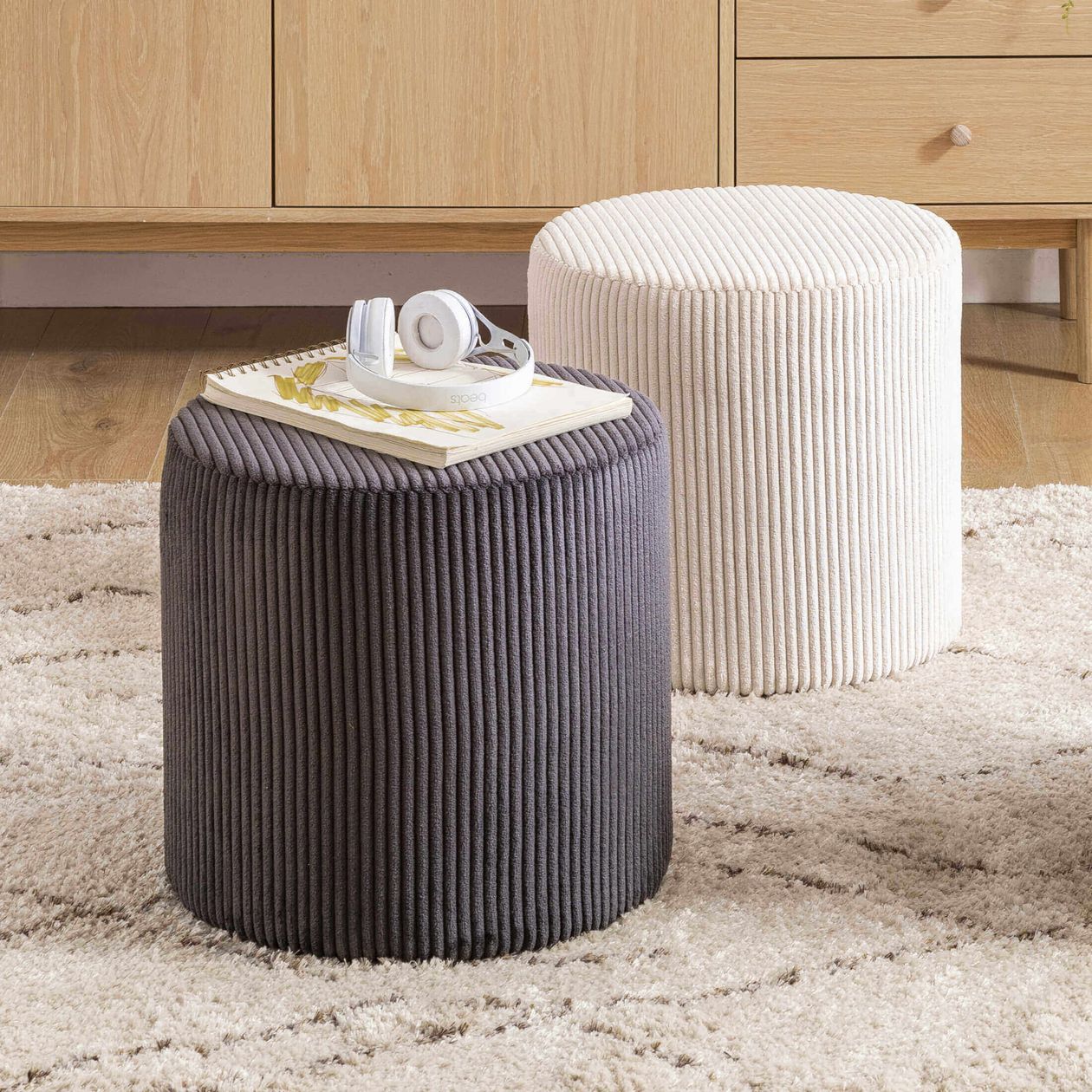Grauer und weißer Pouf aus Cordstoff in einem Raum auf einem Teppich. Auf dem grauen Pouf liegen Kopfhörer und ein Block mit Blättern.