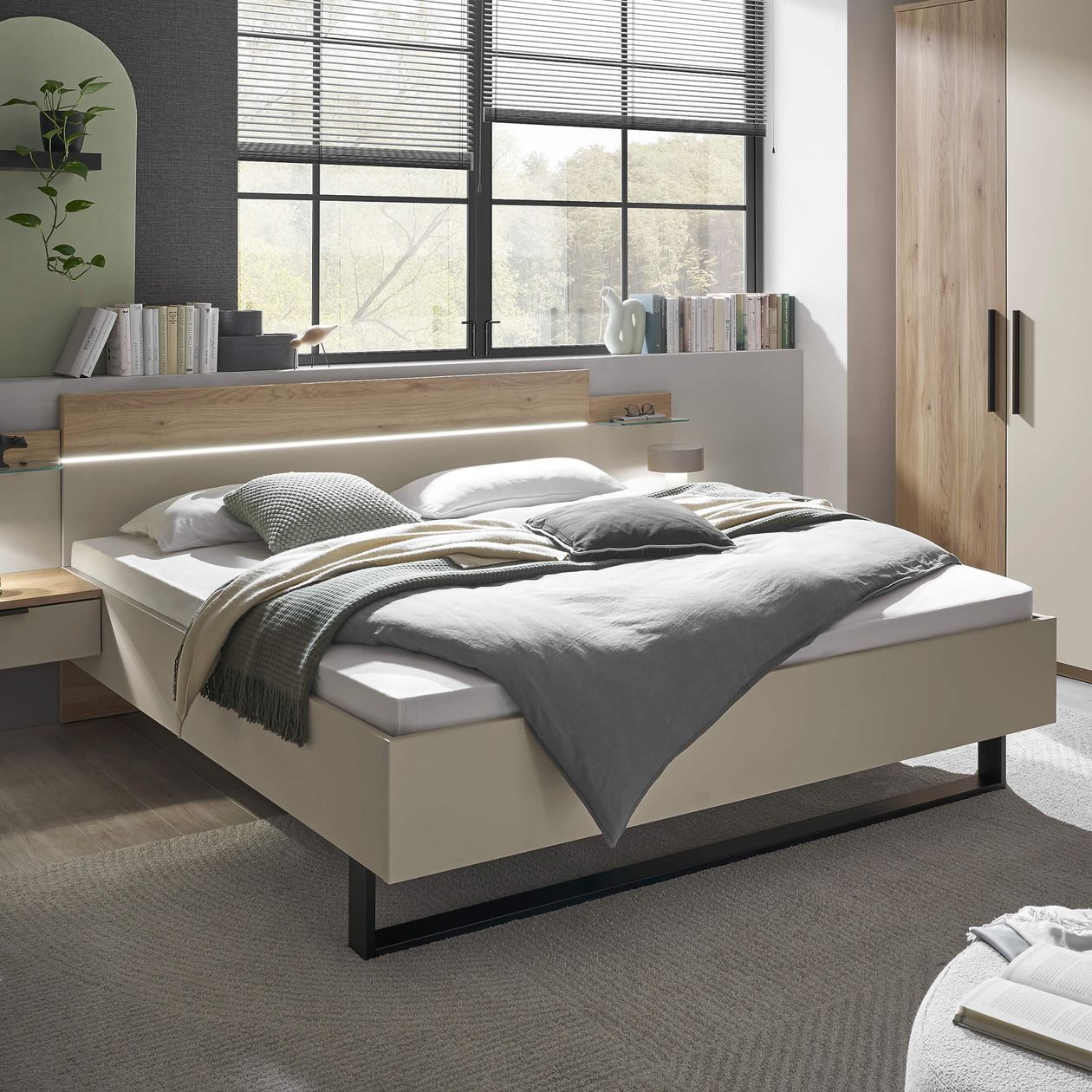 Modernes Schlafzimmer mit großem Fenster, hellem Doppelbett, grauer Bettwäsche, Pflanzen und Büchern im Regal, entspannte Atmosphäre.