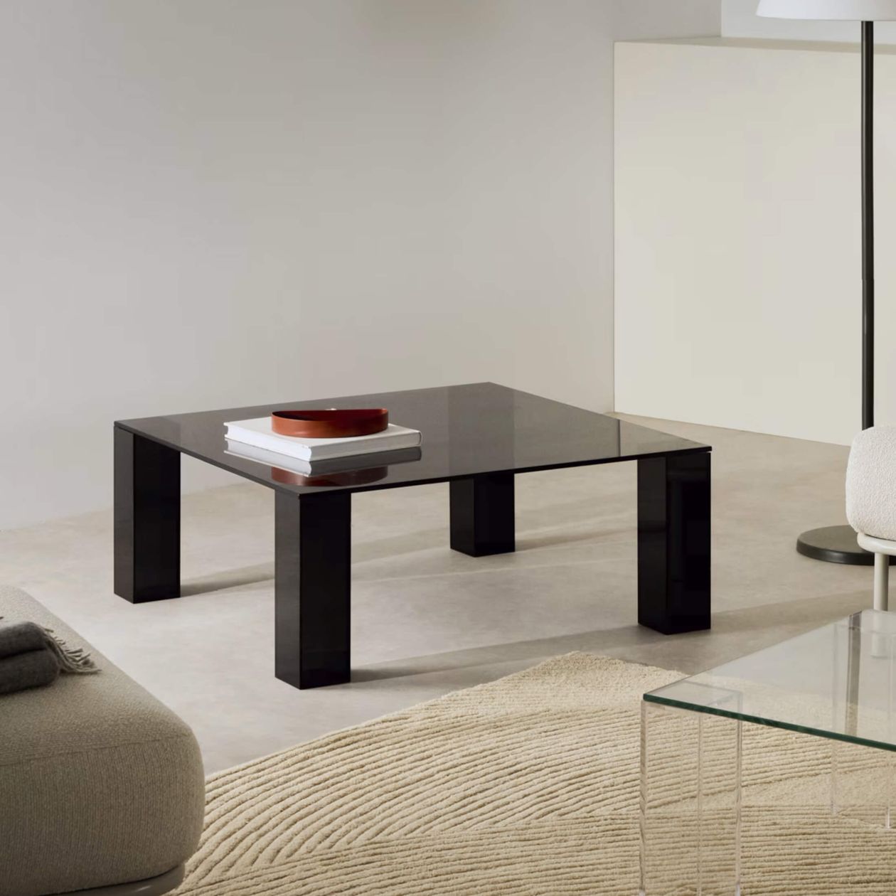 Ein moderner Couchtisch aus schwarzem Glas mit Büchern und einem Tablett, der auf einem beigen, strukturierten Teppich in einem minimalistisch eingerichteten Wohnzimmer steht.
