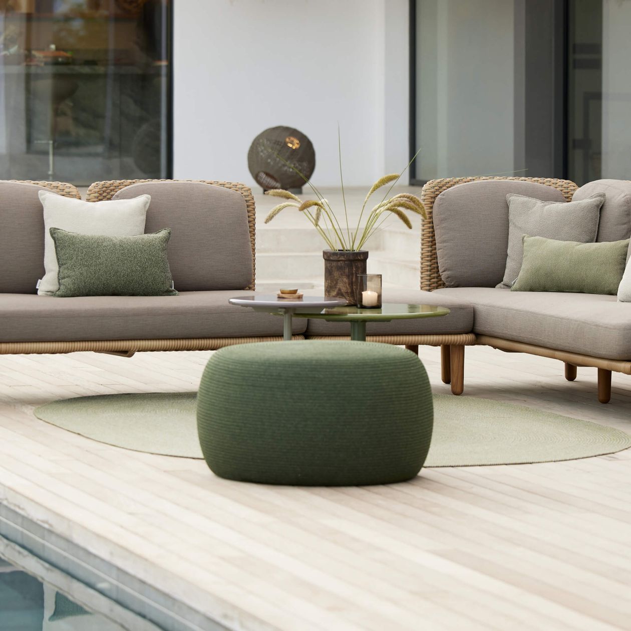 Moderne Terrasse mit grauen Polstermöbeln, grüner Ottomane, rundem Tisch, Pflanzenarrangement und hellem Holzboden neben einem Swimmingpool.