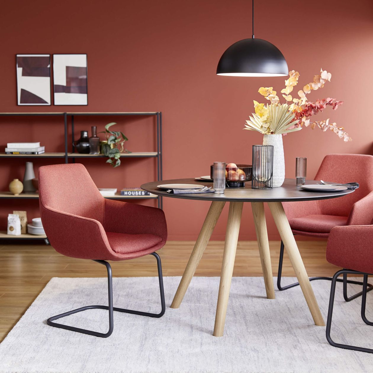 Moderner Essbereich mit runden Tisch, gepolsterten Stühlen in Rot, dekorativer Vase mit Blumen und Regalen vor einer roten Wand.