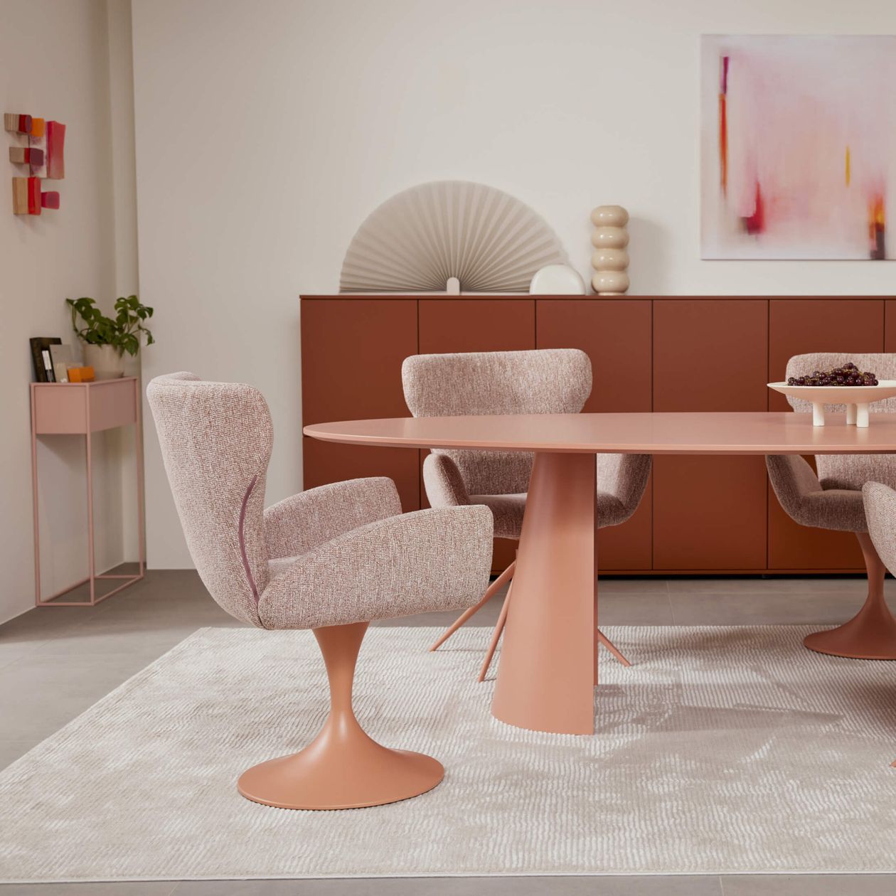 Moderner Essbereich mit ovalem Tisch in Rosa, passenden Stühlen und dekorativer Anrichte. Dezente Kunst an der Wand und Pflanze in der Ecke.