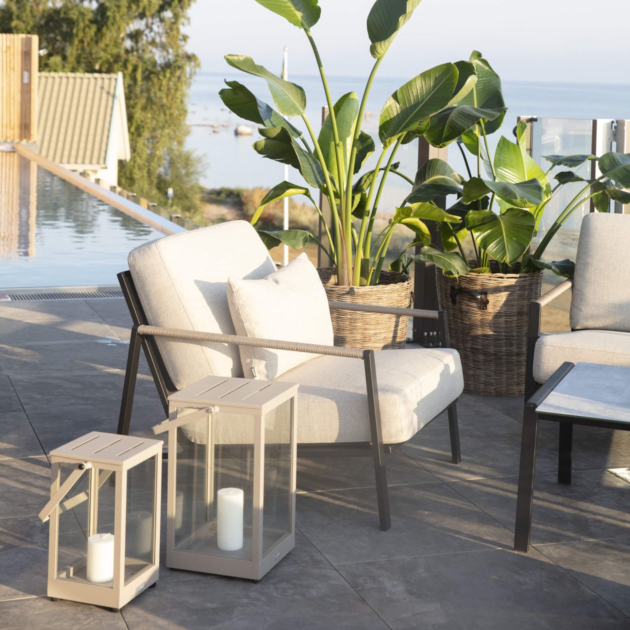 Sonnige Terrasse mit bequemen Stühlen, großen Pflanzen in Körben, Laternen mit Kerzen und Blick auf den Pool und das Meer im Hintergrund.