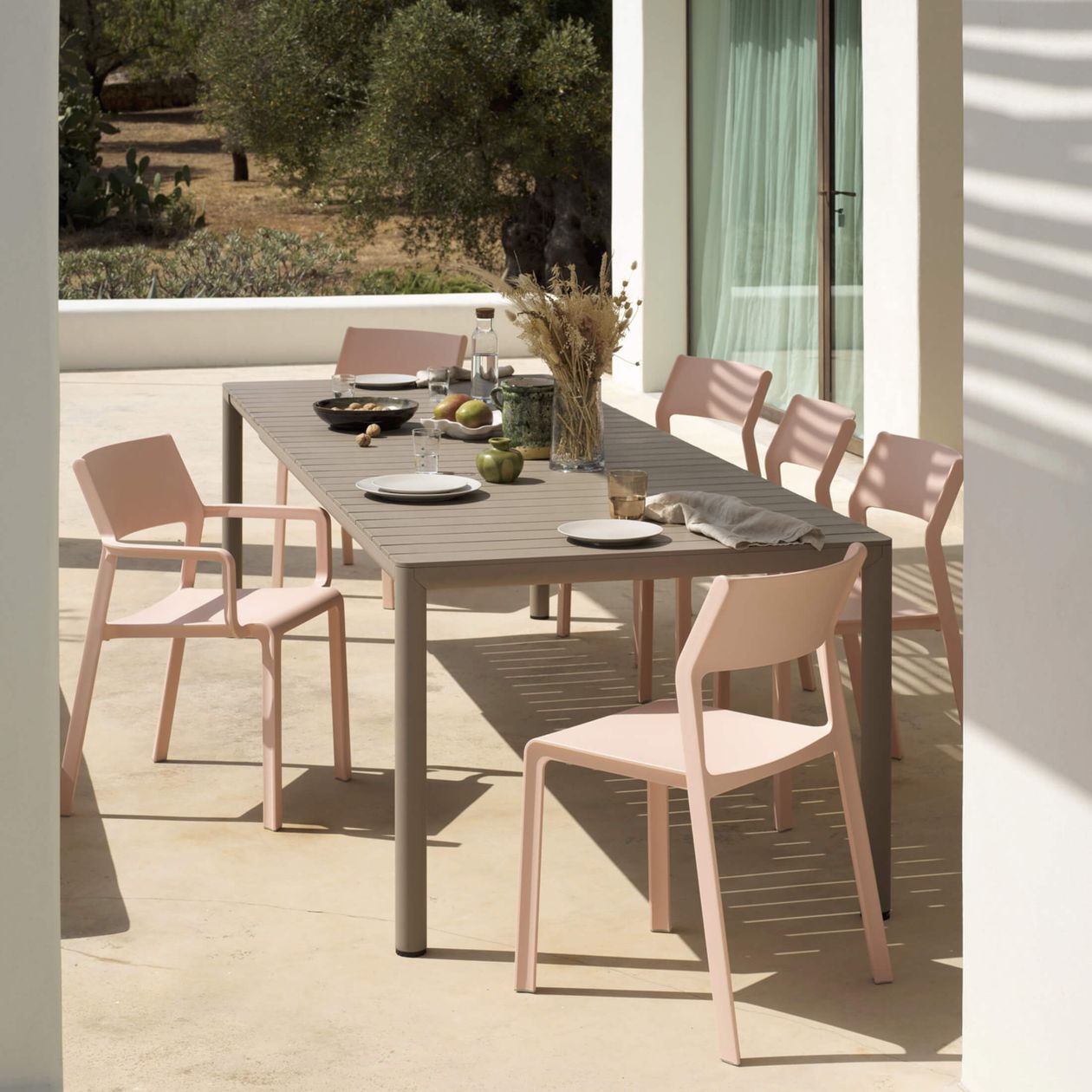 Modernes Terrassen-Esszimmer-Set mit rosa Stühlen und einem Holztisch, gedeckt zum Essen, umgeben von einem sonnigen Blick auf den Garten.