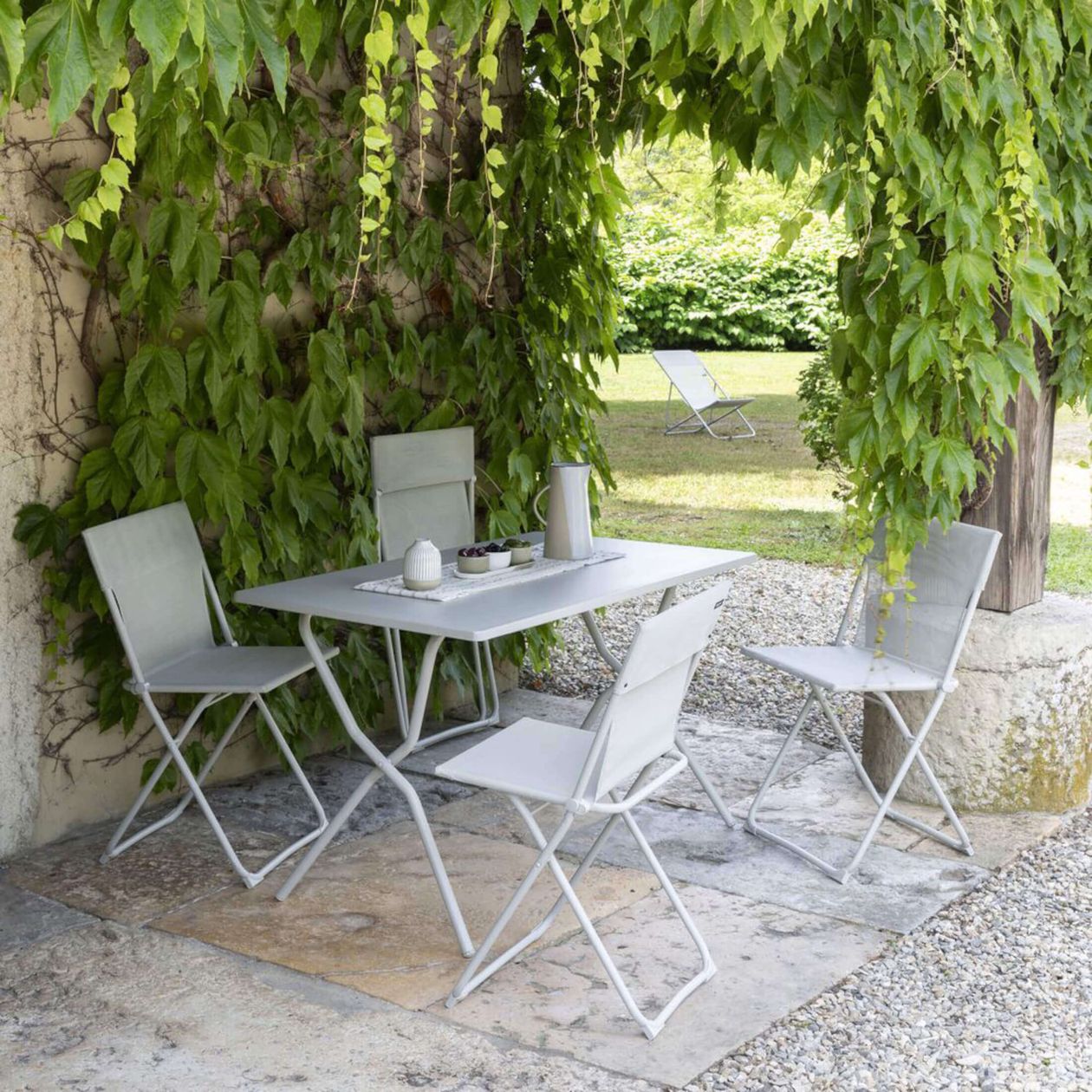 Weiße Gartenstühle und ein Tisch unter einem üppigen grünen Efeubaldachin, aufgestellt auf einer Steinterrasse vor dem Hintergrund eines Gartens und mit einem einzelnen Stuhl auf dem Rasen.