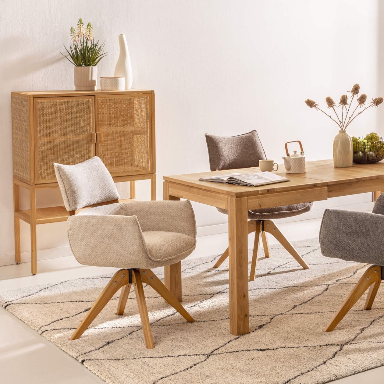 Esszimmer mit Holztisch, gepolsterten Stühlen und Teppich. Links ein Sideboard mit Pflanzen, rechts Vase mit getrockneten Blumen und Keramikgeschirr.