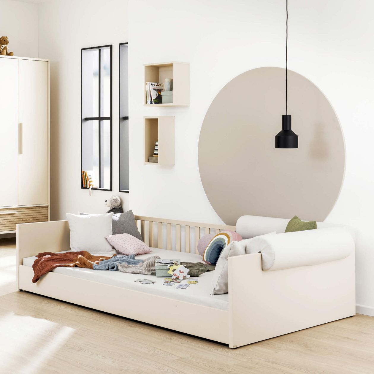 Helles Kinderzimmer mit Tagesbett, Kissen, Decken und einer schwarzen Pendelleuchte. An der Wand befindet sich ein großes, rundes Wandbild.