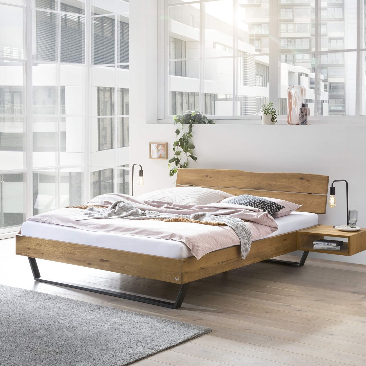 Ein modernes Schlafzimmer mit einem Holzbett auf Podest, rosa-grauer Bettwäsche und einem kleinen Beistelltisch. Im Hintergrund große Fenster mit Blick auf die Stadt.