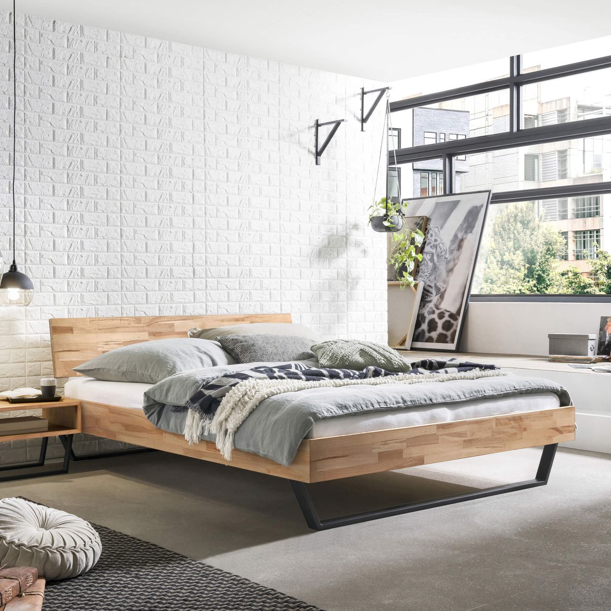 Ein modernes Schlafzimmer mit einem Holzbett, grauer Bettwäsche, einem großen Fenster, einer weißen Backsteinwand und Dekoartikeln. Der Raum ist von Tageslicht durchflutet.