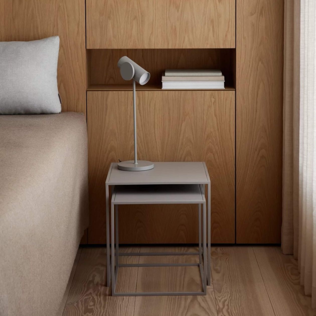 Ein minimalistisches Schlafzimmer mit einem Holzschrank, einer grauen Tischlampe, beiger Bettwäsche und einem Vorhang an der Seite.