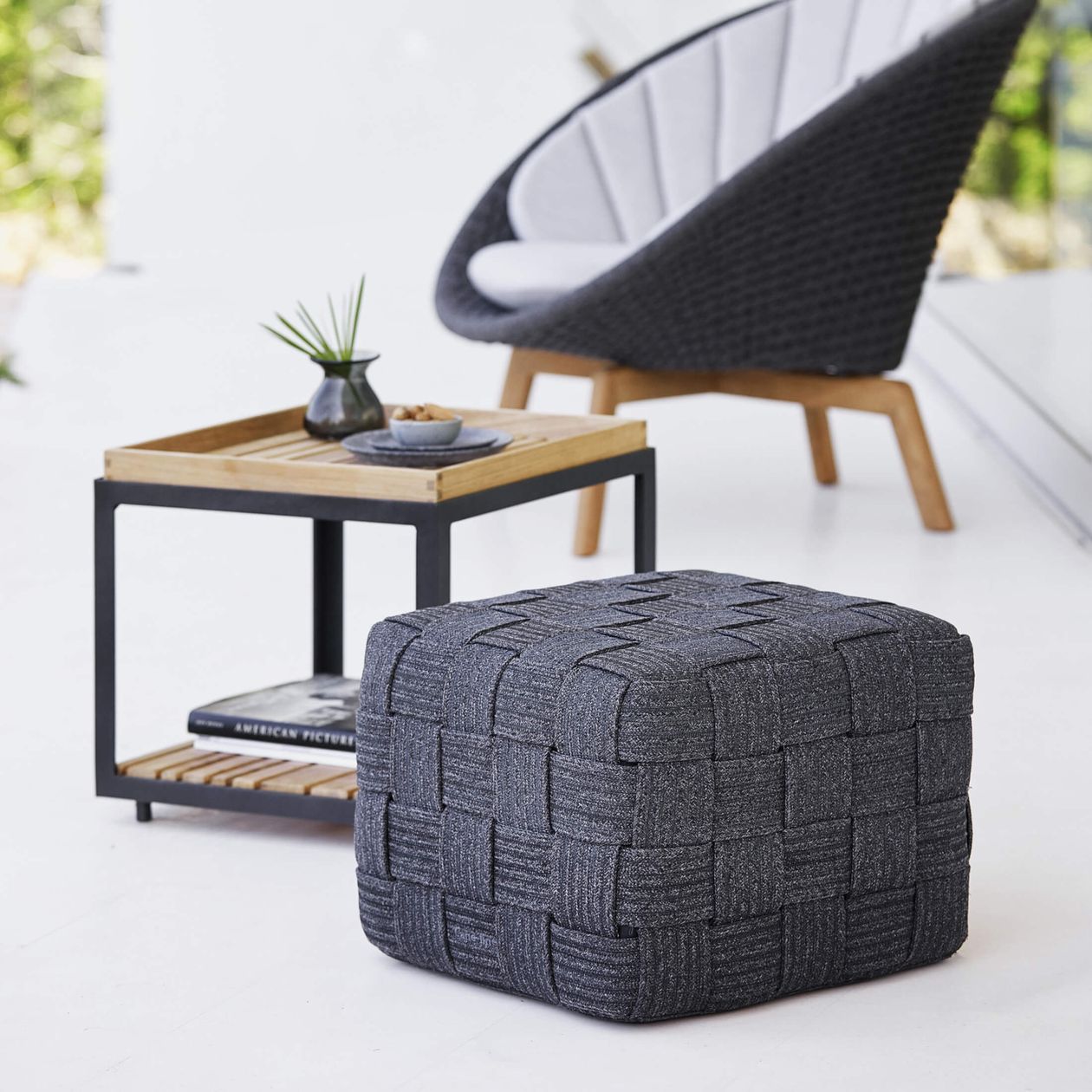 Ein dunkelgrauer, gewebter Pouf neben einem Holztisch mit einer kleinen Vase und einer Schale sowie ein moderner schwarz-weißer Stuhl in einem hellen Raum.