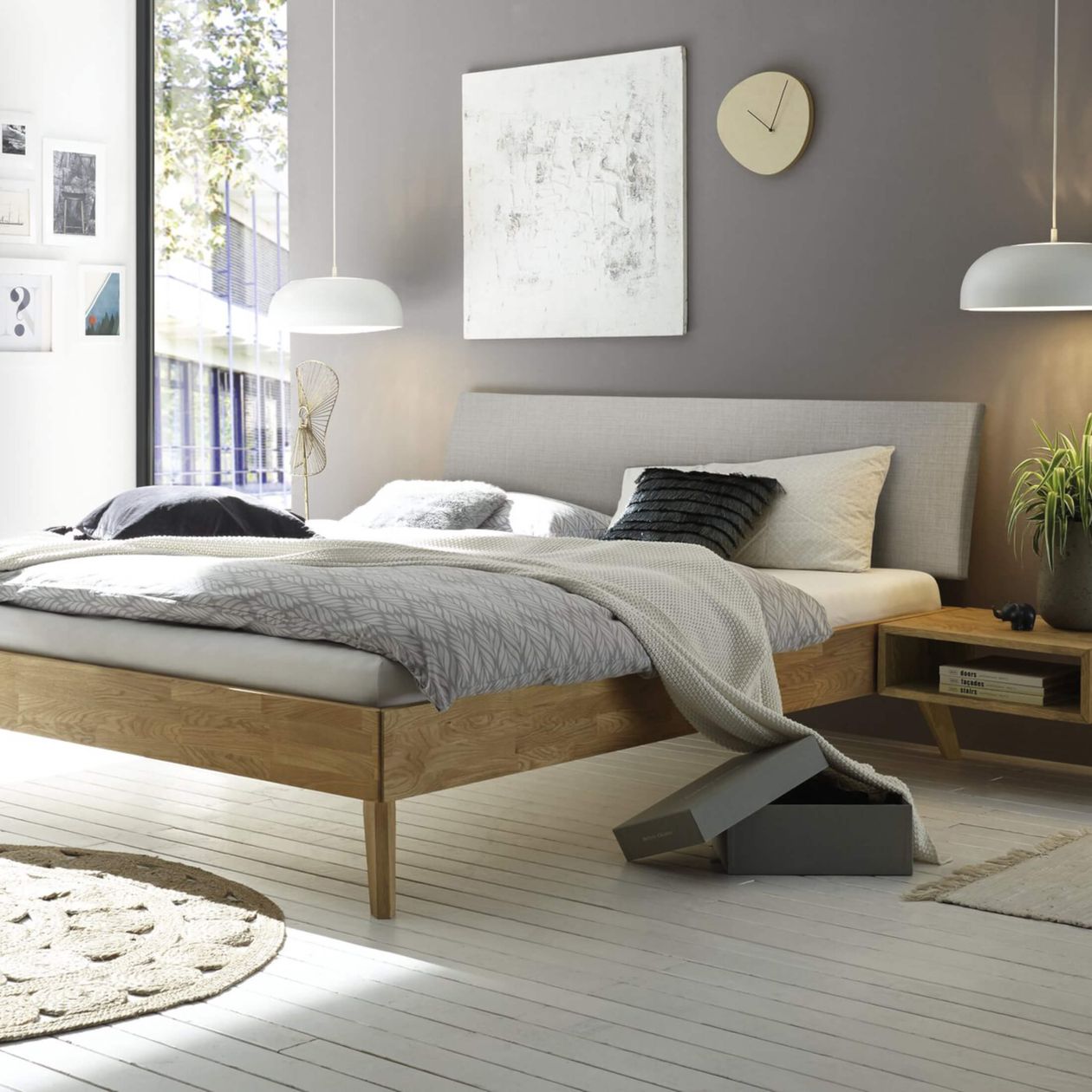 Ein modernes Schlafzimmer mit einem Holzbett, grauer Bettwäsche, Wanddekoration und einer runden Uhr. Über einem kleinen Nachttisch aus Holz hängen Pendelleuchten.