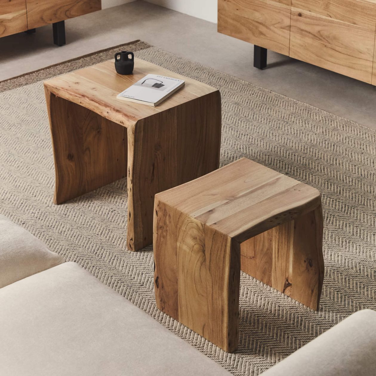 Zwei Beistelltische aus Holz stehen auf einem strukturierten Teppich. Auf einem Tisch liegen ein Buch und eine schwarze Tasse. In einem modernen Wohnzimmer sind teilweise beigefarbene Sofas zu sehen.