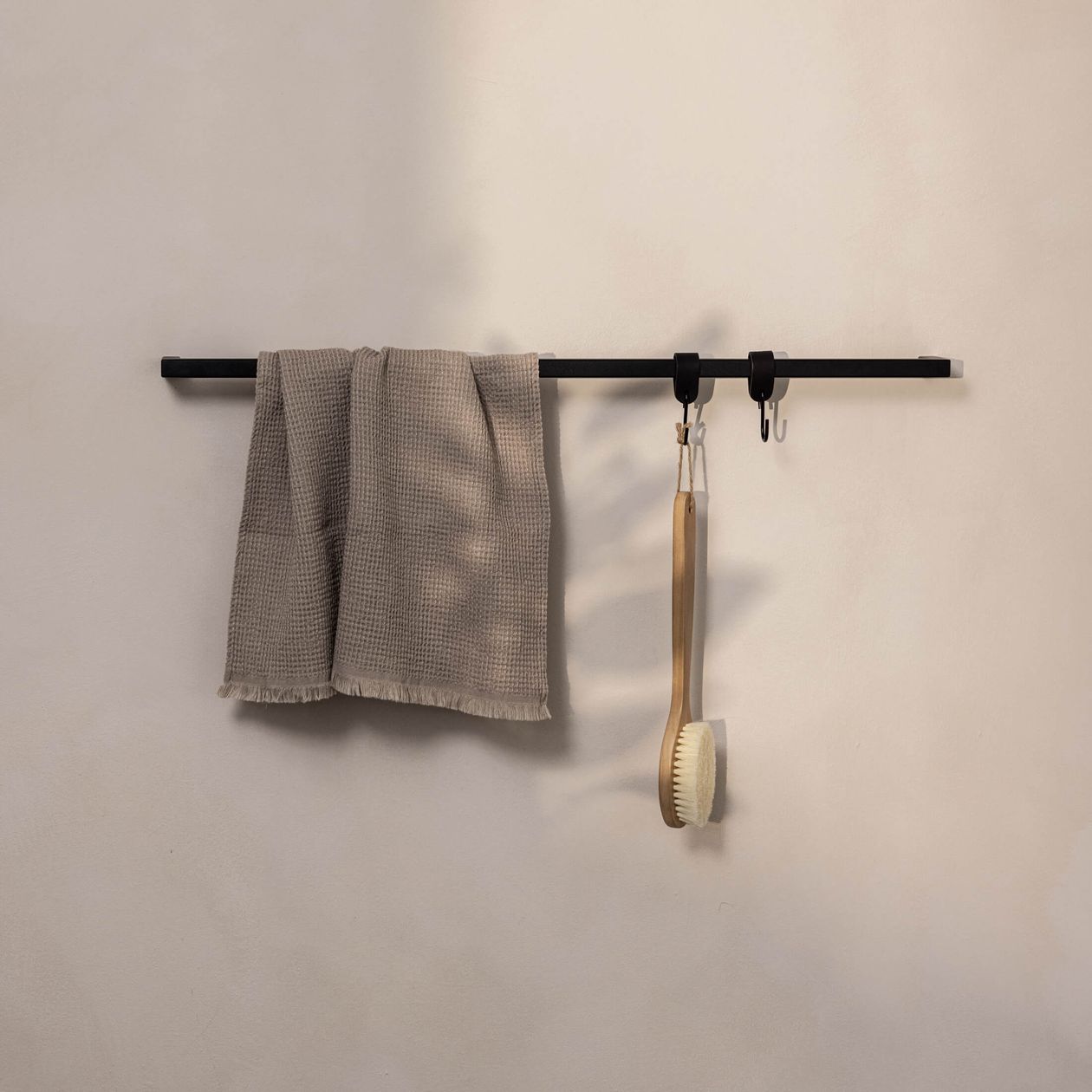 Beigefarbenes Handtuch und Holzborstenbürste hängen an einem schwarzen Wandhaken auf einem minimalistischen, hellen Hintergrund.