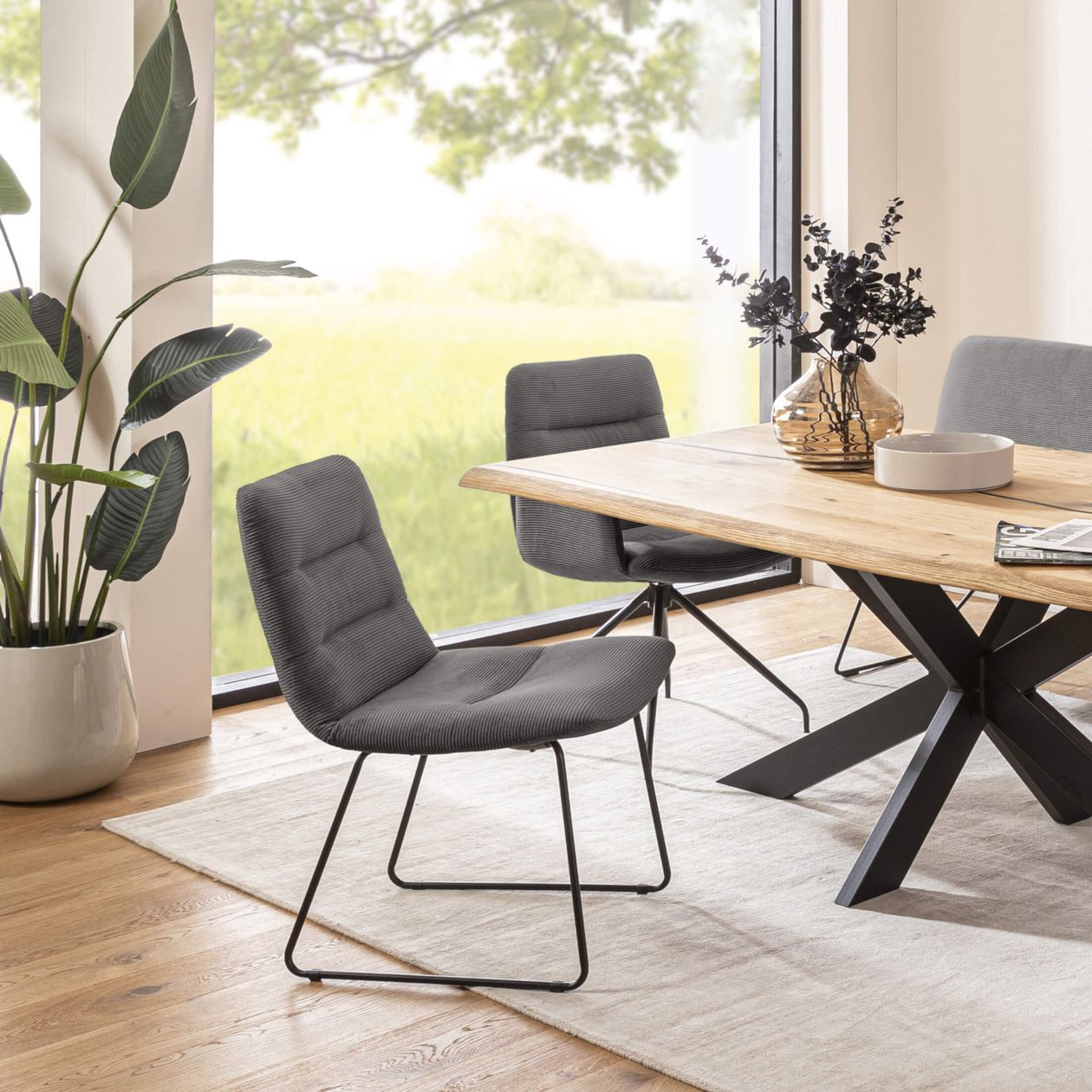Esszimmer mit Holztisch, grauen Stühlen und einem Teppich. Große Fenster bieten Blick auf grüne Wiese. Zimmerpflanze und Vase zur Dekoration.
