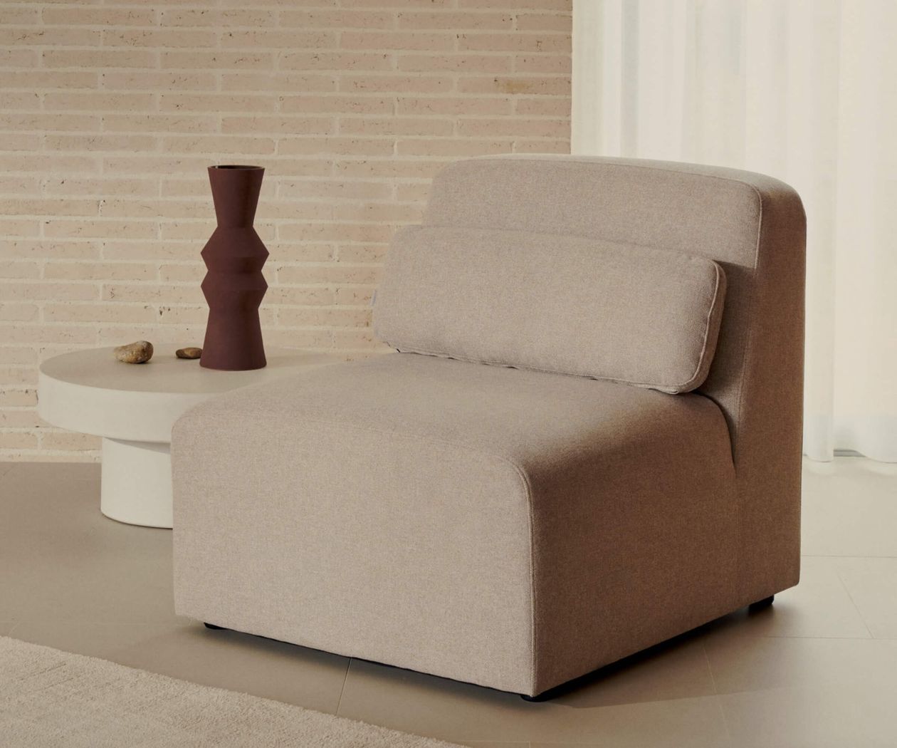 Beiges Ecksofa vor einer Ziegelwand, neben einem runden weißen Tisch mit brauner Vase und kleinen Steinen darauf.
