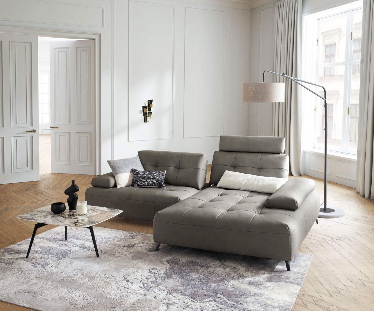 Elegantes Wohnzimmer mit grauem Ledersofa, Marmortisch, Stehlampe und großen Fenstern. Helle Wände und Holzboden schaffen eine moderne Atmosphäre.
