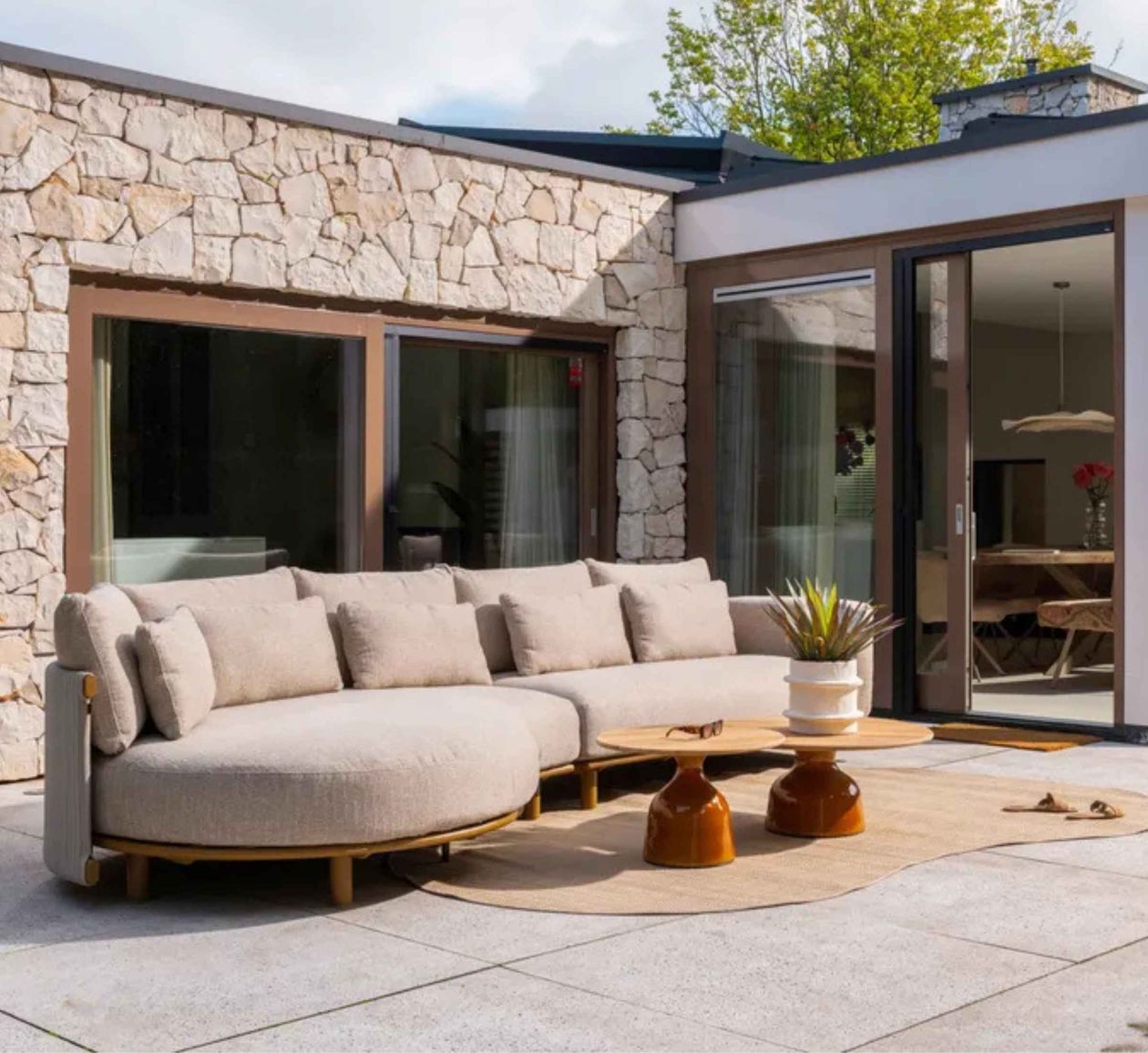 Moderner Patio mit beigefarbenem Sofa, Holzcouchtischen und Topfpflanze. Steinwand und große Fenster im Hintergrund.
